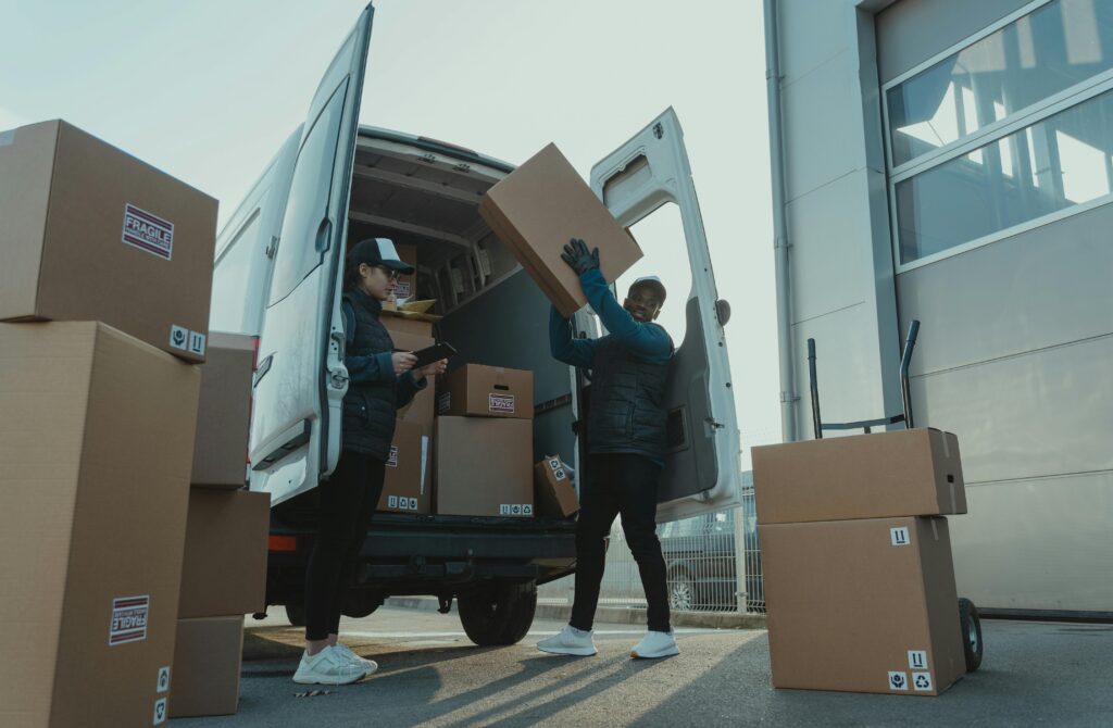 Workers loading boxes into a delivery van at a warehouse facility outdoors.